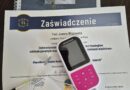Zaświadczenie o ukończeniu kursu logopedycznego z elektrostymulacji leżące na biurku, obok bindowanego podręcznika i przenośnego elektrostymulatora Logotens z podłączonymi przewodami.