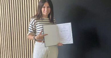 Uśmiechnięta kobieta w bluzce w paski i jasnych spodniach trzyma otwartą teczkę z zaświadczeniem, wskazując na nią palcem, na tle nowoczesnej ściany z drewnianymi lamelami.