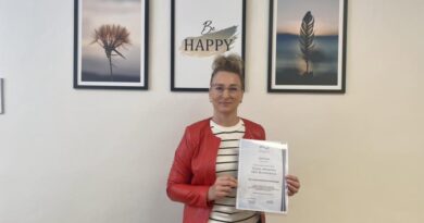 Kobieta w okularach i czerwonej kurtce trzyma w rękach certyfikat szkoleniowy, stojąc na tle jasnej ściany, na której wiszą trzy obrazy w czarnych ramkach.