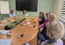 Grupa kobiet siedzi wokół dużego, owalnego stołu konferencyjnego podczas spotkania w sali szkoleniowej z dużym ekranem na ścianie.