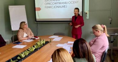 Kobieta w bordowym garniturze prowadzi prezentację dla grupy osób siedzących przy dużym stole konferencyjnym. Na ekranie za nią widnieje napis: „ZADANIA PEDAGOGA SZKOLNEGO W OPRACOWYWANIU OCENY FUNKCJONALNEJ”.