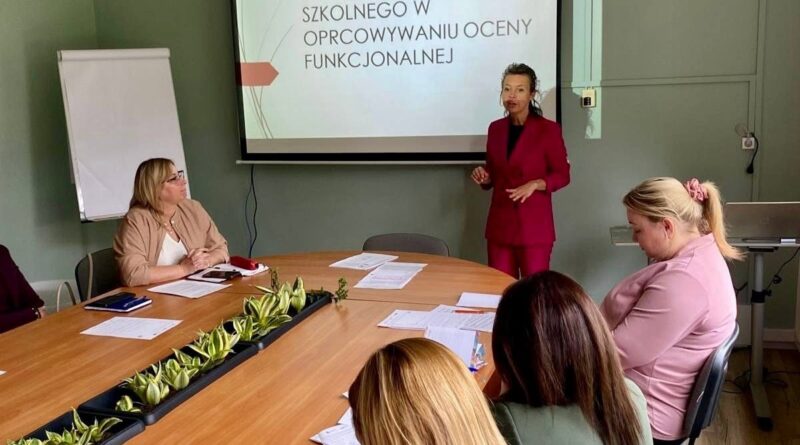 Kobieta w bordowym garniturze prowadzi prezentację dla grupy osób siedzących przy dużym stole konferencyjnym. Na ekranie za nią widnieje napis: „ZADANIA PEDAGOGA SZKOLNEGO W OPRACOWYWANIU OCENY FUNKCJONALNEJ”.